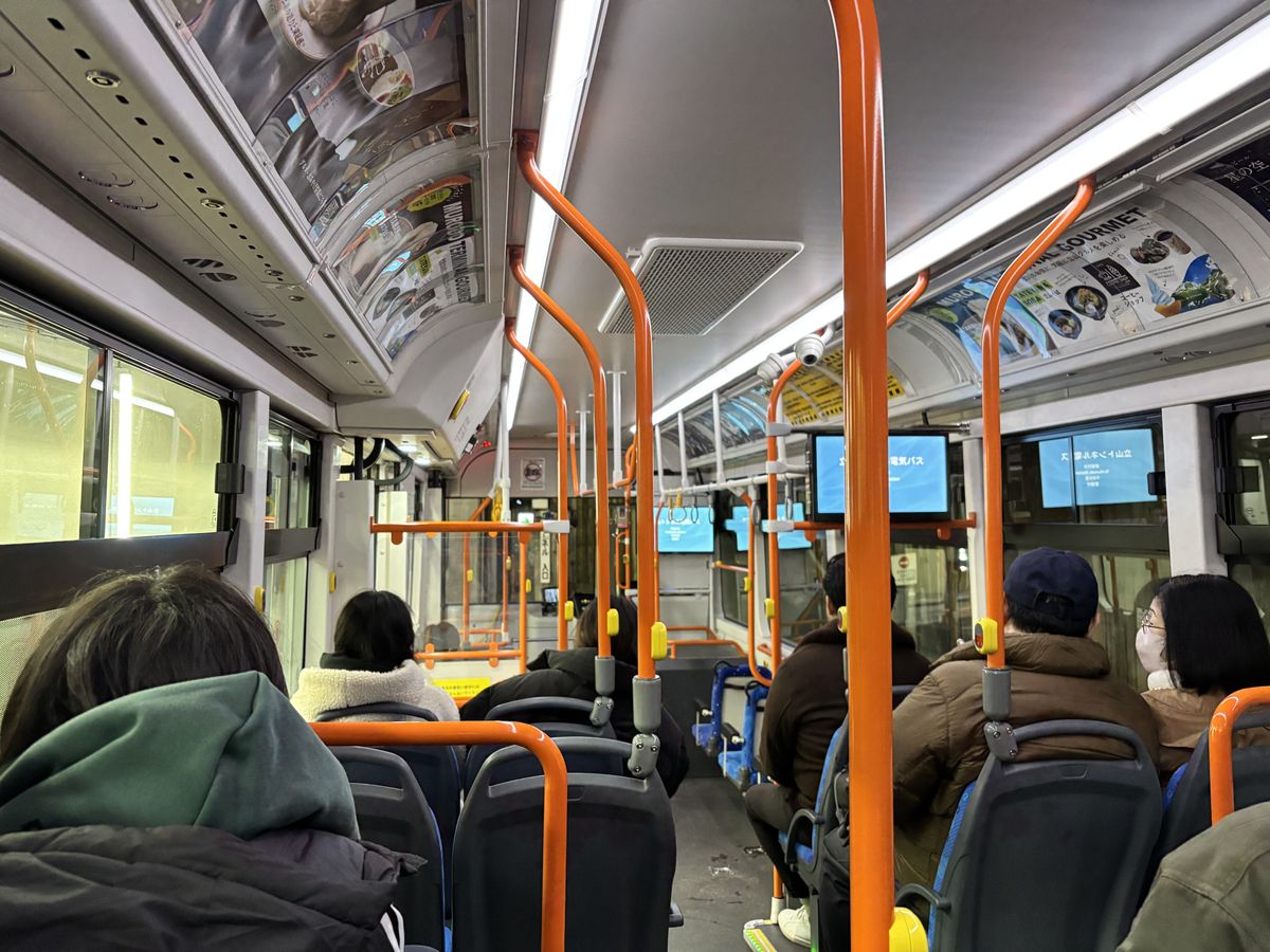 Warmly dressed passengers aboard a highland bus on the Tateyama Kurobe Alpine Route, orange grab poles lining the aisle