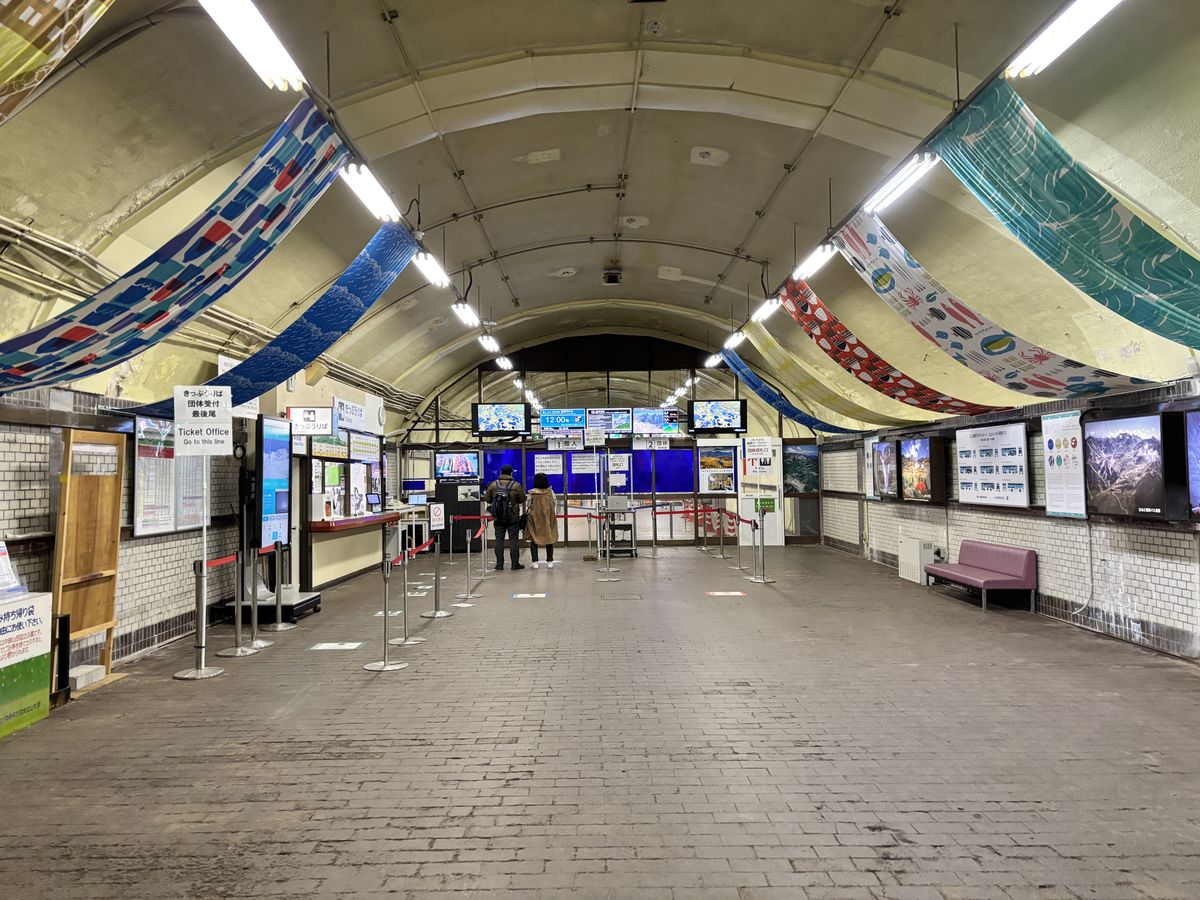 Two travellers walking toward the ticket gate through the arched tunnel concourse of an Alpine Route station