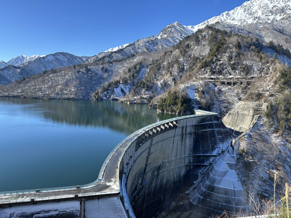 Winter panorama from the dam crest walk: the curving dam wall and lake framed by the snowy Japanese Alps