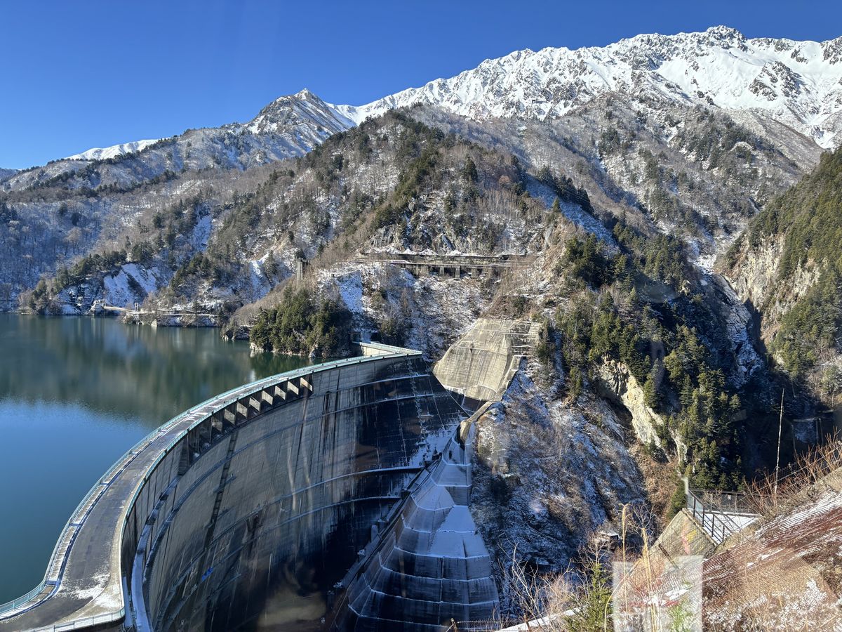 View from the dam walk of the huge arched wall holding back blue-green water, snow-capped peaks behind