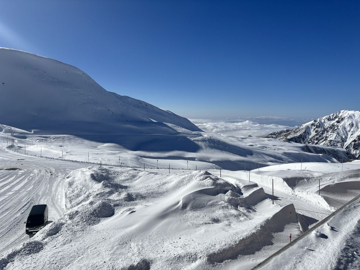 Gondola lift rising over snowy slopes below the Tateyama mountain range under a wide blue sky