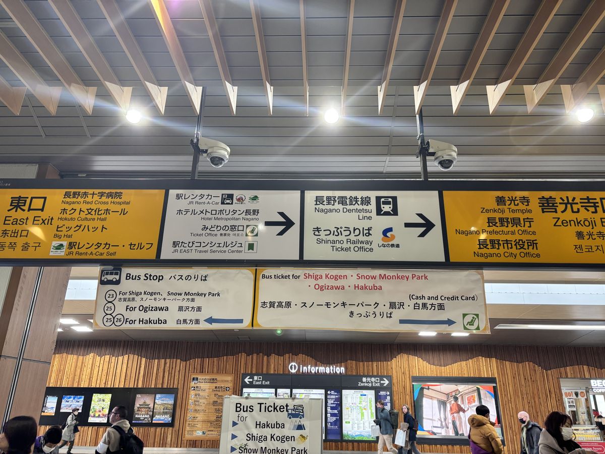 Travellers passing beneath signs for the east and Zenkoji exits and the bus stops inside Nagano Station