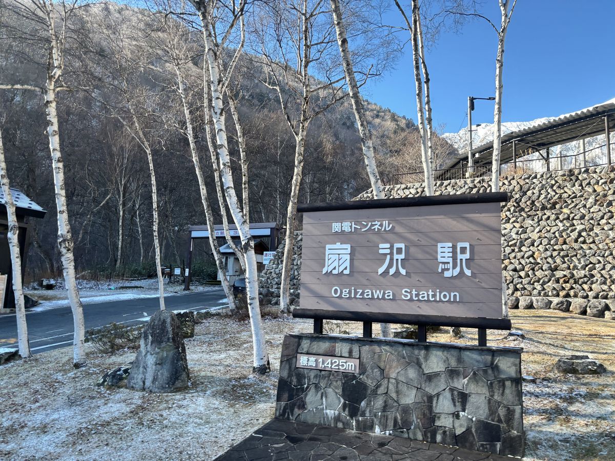 Birch trees lined up against snowy mountains, with the name sign of Ogizawa Station standing in front