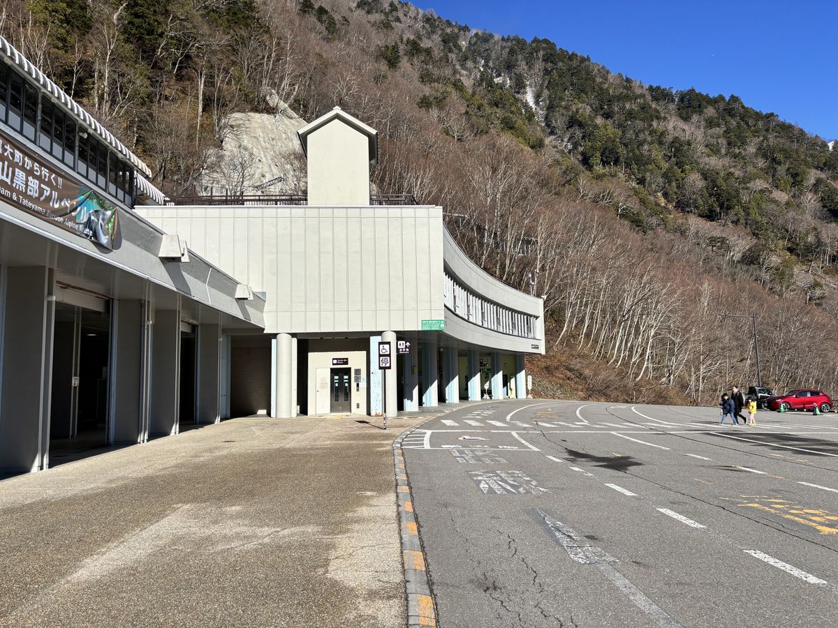 Parent and child crossing the parking lot by the white terminal building at the foot of the Tateyama Kurobe Alpine Route
