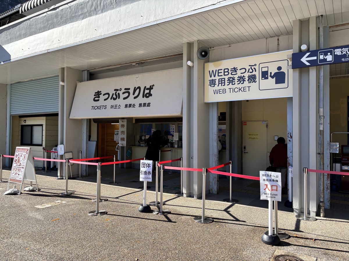 Ticket window marked Kippu Uriba at Ogizawa, red belt barriers lining up travellers for the Kurobe Dam bus